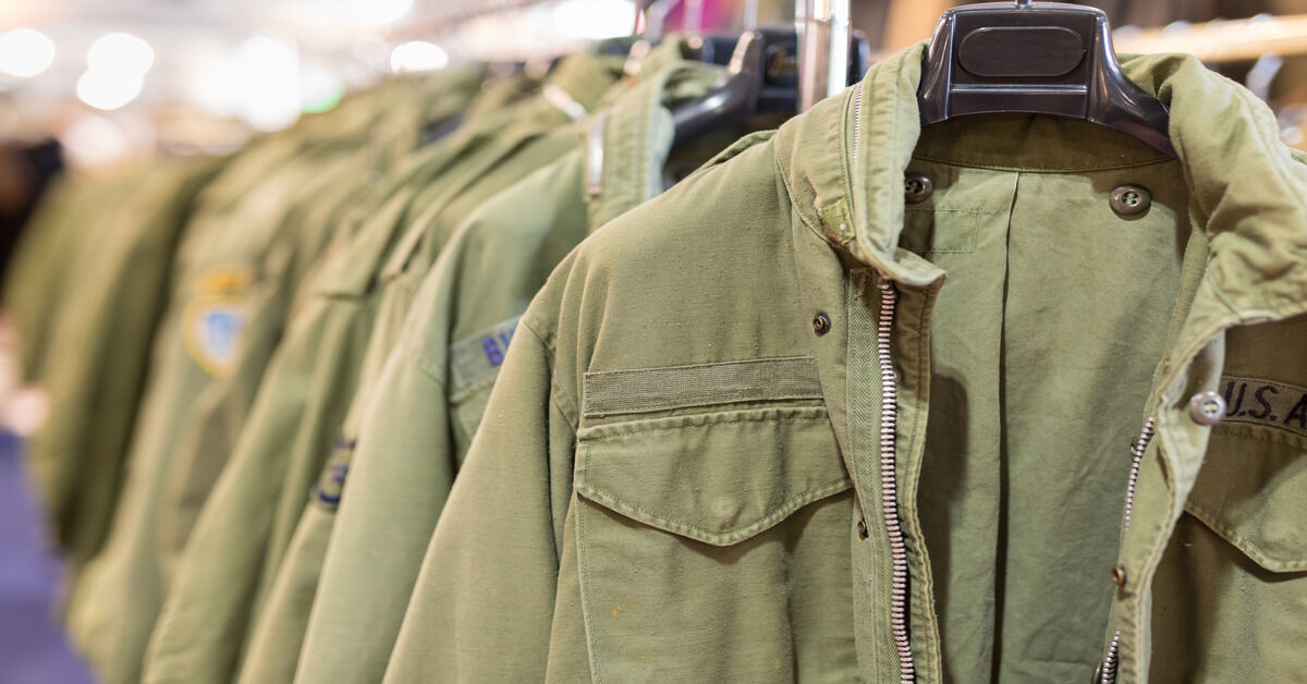 A close-up of a green military jacket with the letters U.S.A. on the chest. Behind is a rack of more of the same jackets.